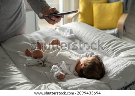 Unknown woman caucasian mother using mobile phone taking photos of her newborn baby boy or girl infant lying on the bed at home in day - modern motherhood and growing up concept