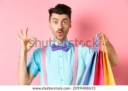Funny guy in bow-tie showing OK sign and shopping bags, praising good deal in shops, standing over pink background