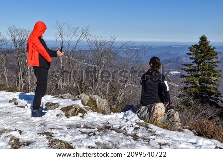 A woman sits on a stone and a man photographs her on his phone. Snow, mountain two people are photographed in nature. Beautiful winter landscape, mountains and trees. A loving couple takes photo shots