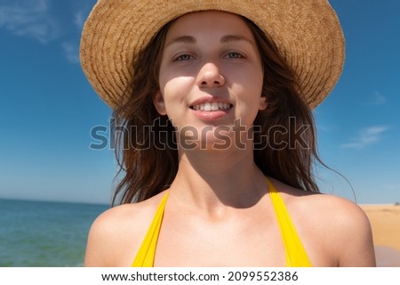 Close-up portrait of attractive caucasian young woman in straw hat and yellow swimsuit smiling against blue sky and sea on a sunny day
