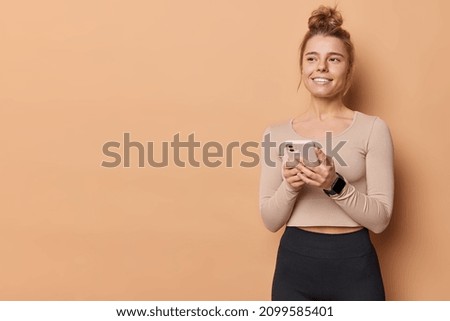 Sporty slim European woman holds mobile phone for messaging and surfing internet dressed in sportswear has satisfied dreamy expression poses against beige background blank copy space for text