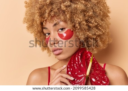 Serious woman takes care of skin applies hydrogel patches under eyes to reduce wrinkles holds red calla flower looks directly at camera isolated over beige background. Beauty and wellness concept