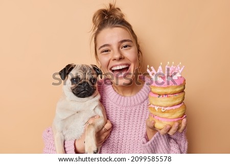 Joyful happy woman enjoys birthday party celebration poses with pug dog and delicious festive donuts wears knitted sweater laughs out gladfully isolated over beige background. Pet anniversary Royalty-Free Stock Photo #2099585578
