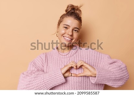 Lovely affectionate young European woman with toothy smile makes heart gesture confesses in love wears knitted sweater isolated over beige background expresses romantic feelings. Be my valentine