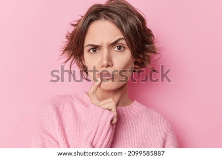 Serious skeptical woman keeps index finger near folded lips raises eyebrows suspiciously has doubts suspects someone tries to search solution wears casual jumper isolated over pink background Royalty-Free Stock Photo #2099585887