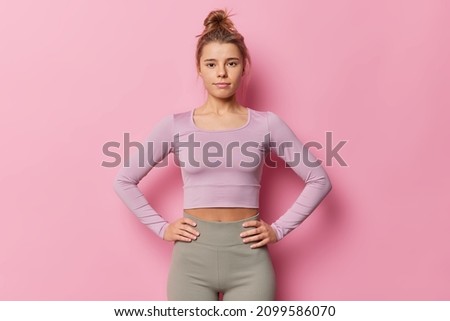 Indoor shot of serious sporty woman keeps hands on waist dressed in tracksuit looks attentively at camera has combed hair leads active lifestyle poses against pink background. Sporty lifestyle