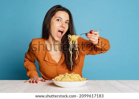 Looks very tasty. Beautiful young girl eating delicious Italian pasta isolated on blue studio background. Holidays, traditions, food, popularity, cafe, love. Healthy carbohydrates. Copy space for ad Royalty-Free Stock Photo #2099617183