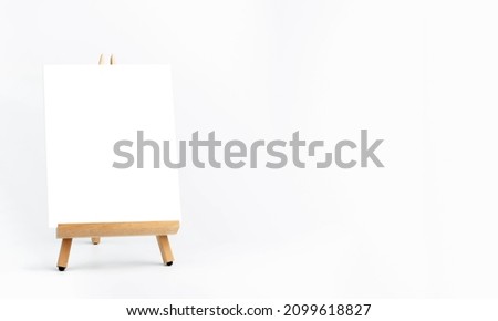 White blank artist frame on a small wooden easel on white background with copy space. Advertising mockup artboard for pictures or artwork. Painting frame template on easel stands display banner. Royalty-Free Stock Photo #2099618827