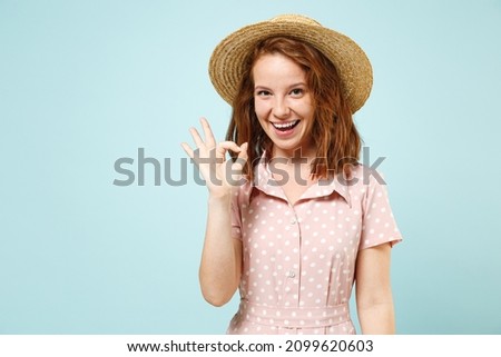 Satisfied smiling young redhead curly woman 20s show ok okay wears casual pink dress straw hat look camera isolated on pastel blue color background studio portrait. People emotions lifestyle concept