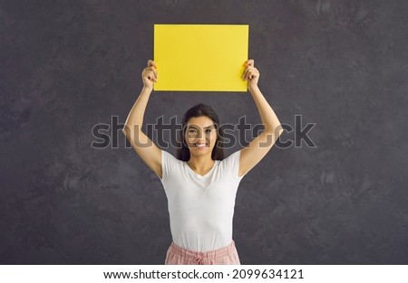 Positive joyful woman standing on gray background holding blank yellow sheet of paper for text. Brunette woman with beautiful smile holds sheet of paper over her head. Copy space. Advertising banner.