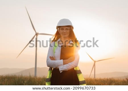 Beautiful caucasian woman in white helmet working with digital tablet at renewable energy farm. Female inspector controlling functioning of wind turbines outdoors. Royalty-Free Stock Photo #2099670733