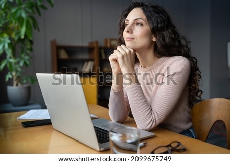 Dreamy businesswoman freelance writer looking in window, distracted from laptop, sitting at work desk in office. Happy young female freelancer thinking of new opportunity, project strategy or vacation Royalty-Free Stock Photo #2099675749