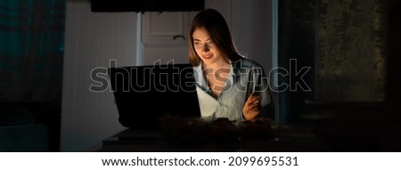 A lonely woman in casual clothes sits at the table at home at night and watches online TV with a smile on her face. Baner. copy space.