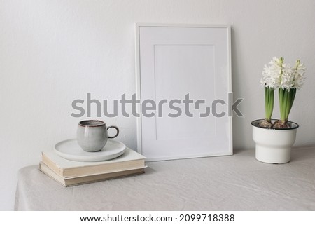 Spring breakfast still life scene. Cup of coffee, books. Empty white picture frame mockup. Beige linen tablecloth. Potted white hyacinth plant. Farmhouse, Scandinavian interior. Easter holiday concept
