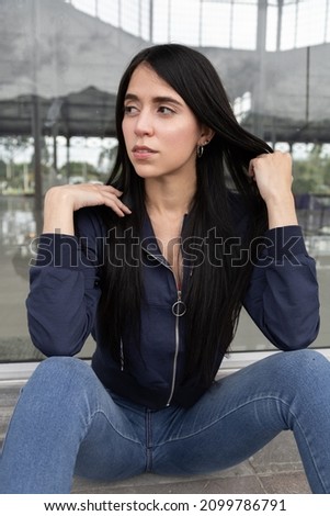 young latin woman with long straight black hair posing sitting in casual jacket and jeans, urban lifestyle and beauty in the city