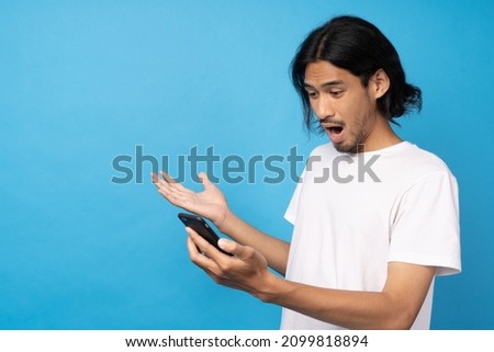 Cool asian man excited and surprised by smartphone on blue background. A man saw a sale on an online shopping website or See the results of the football team cheering competition.