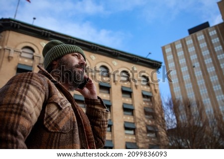 A bearded man wearing a green knitted beanie uses a smartphone in the city in this technology lifestyle concept.  The cell phone uses data and a wireless network as he communicates digitally.