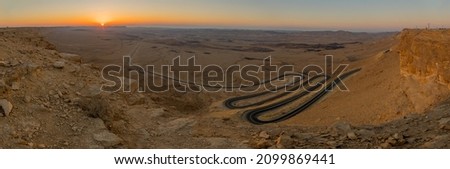 Panoramic sunrise view of a winding road and the landscape of Makhtesh (crater) Ramon, in the Negev Desert, Southern Israel. It is a geological landform of a large erosion cirque Royalty-Free Stock Photo #2099869441