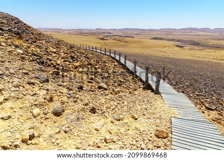 View of an elevated footpath and hexagonal rock formations in the Carpentry site, part of Makhtesh (crater) Ramon, in the Negev Desert, Southern Israel Royalty-Free Stock Photo #2099869468