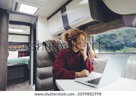 Adult woman use laptop computer inside a camper van recreational vehicle sitting at the table with bedroom in background and nature park outside the windows. Concept of travel and remote worker people Royalty-Free Stock Photo #2099882998