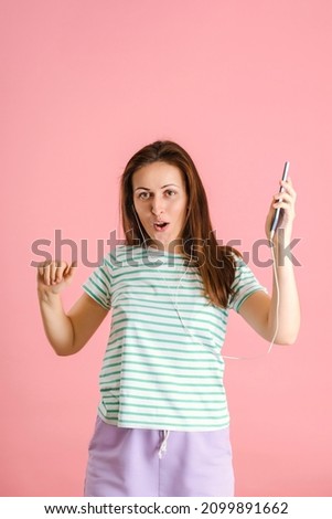 A charming middle-aged brunette dances and sings while listening to music through headphones from a smartphone. A positive girl dances to music on a pink background.