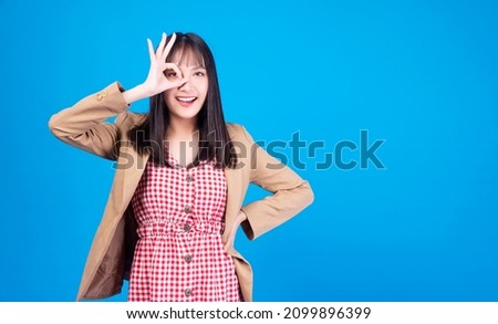 Confident cheerful Asian businesswoman wear brown jacket using finger make sign ok cover on own eyes emotion good-looking or vision or approve while standing over isolated blue background.