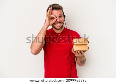 Young caucasian delivery man holding a burger isolated on white background excited keeping ok gesture on eye.