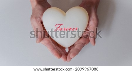 A man's hands holding a white heart. White background. Red lettering in italics "Forever". Concept of love and valentines day.