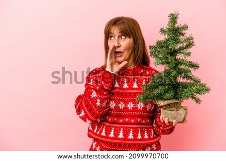 Middle age caucasian woman holding Christmas tree isolated on pink background is saying a secret hot braking news and looking aside