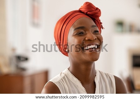 Smiling middle aged african american woman with orange headscarf. Beautiful black woman in casual clothing with traditional turban at home laughing. Portrait of mature carefree lady looking away. Royalty-Free Stock Photo #2099982595
