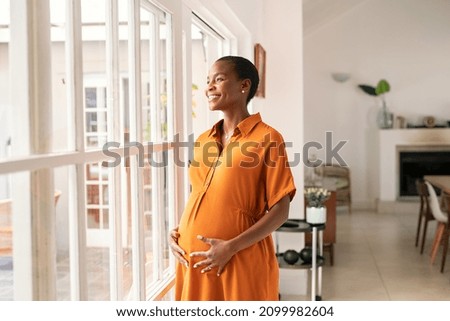 Mid black pregnant woman standing near window at home and thinking about her future family. Smiling african american lady with hands on belly imagine the growth of his baby. Middle aged pregnant woman Royalty-Free Stock Photo #2099982604