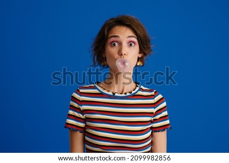 White surprised woman in rainbow earrings making bubble gum isolated over blue background
