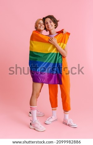 Excited lesbian couple hugging while posing with rainbow flag isolated over pink background