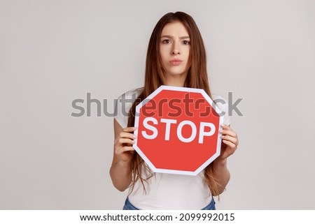 Portrait of strict woman standing with serious facial expression, showing red stop symbol, looking at camera, wearing white T-shirt. Indoor studio shot isolated on gray background.