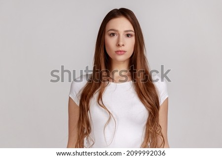 Portrait of strict attractive woman standing looking at camera, feels confident focused self-assured, expressing seriousness, wearing white T-shirt. Indoor studio shot isolated on gray background. Royalty-Free Stock Photo #2099992036