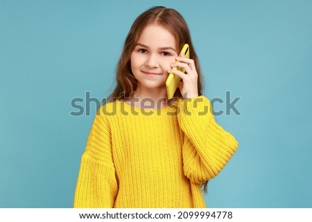 Cute little girl calling on cell phone, good cellular, comfortable to use children mobile device, wearing yellow casual style sweater. Indoor studio shot isolated on blue background.
