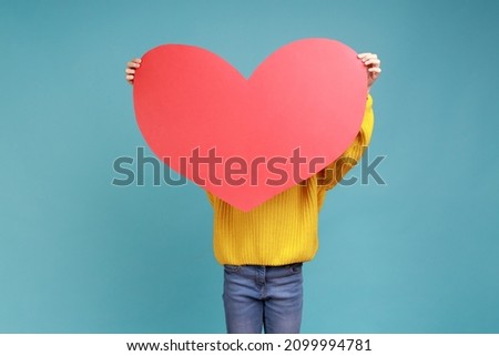 Portrait of anonymous little girl hiding behind big red heart, holding symbol of love and affection, wearing yellow casual style sweater. Indoor studio shot isolated on blue background.