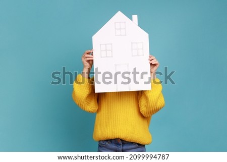 Portrait of unknown little kid hiding behind paper house, affordable housing program, advertisement, wearing yellow casual style sweater. Indoor studio shot isolated on blue background. Royalty-Free Stock Photo #2099994787