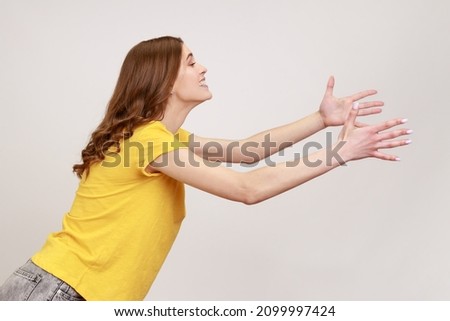 Side view of kind young female with brown hair outstretching hands as if giving for free, offering to embrace, complacency and egoism concept. Indoor studio shot isolated on gray background. Royalty-Free Stock Photo #2099997424