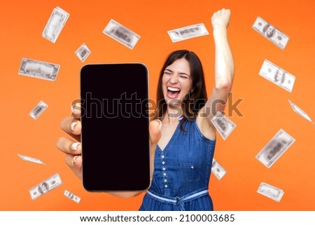Online investment, winner overjoyed rich woman and money earning. Portrait of amazed young woman showing mobile empty display and celebrating her victory. indoor isolated on orange background. Royalty-Free Stock Photo #2100003685