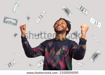 Money rain, winner and rich. Hurray. satisfied happy man with dreadlocks rising hands up with toothy smile on face, pleased, amazed with money falling. Indoor studio shot isolated on gray background