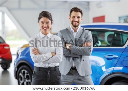 Smiling friendly car seller standing in car salon with arms crossed. It's always a pleasure for both sides to buy a car on a right place. Royalty-Free Stock Photo #2100016768