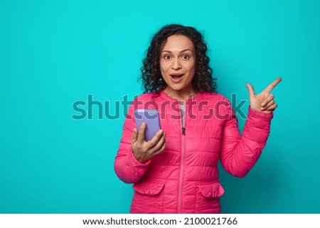 Happy curly haired brunette, beautiful African woman in pink jacket, with wide open eyes in surprise looking at camera holding mobile phone and pointing with finger on blue background with copy space