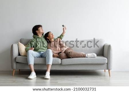Split system. Happy young Asian couple with remote relaxing under air conditioner, sitting on couch at home, mockup for design. Happy millennial spouses using climate control indoors Royalty-Free Stock Photo #2100032731