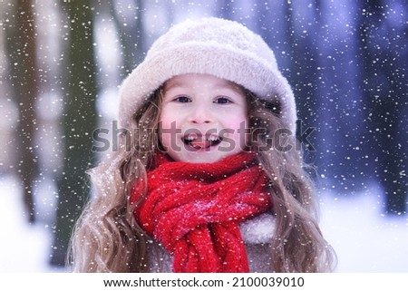 Portrait of beautiful cute baby kid, happy positive cheerful joyful child girl catches snowflakes outdoors in winter snowy park, sticking tongue out, smiling in warm clothes, hat, scarf at snowy day