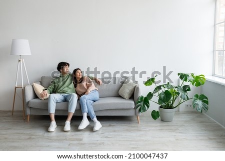 Spending weekend together. Happy millennial Asian couple resting on couch at home, empty space. Cheerful young spouses relaxing and enjoying each other's company in living room Royalty-Free Stock Photo #2100047437