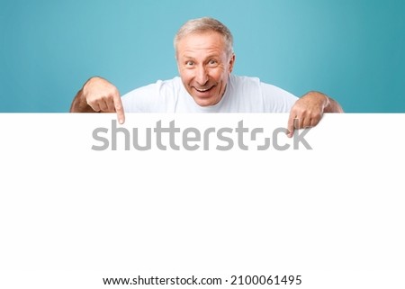 Great Offer. Happy Excited Mature Man Peeping Out Of White Advertisement Board, Pointing Fingers Down At Free Space For Your Text Or Design. Smiling Cheerful Guy Showing Mockup, Standing On Blue Wall