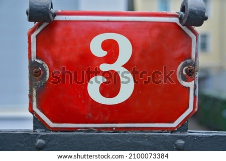 A red house number plaque, showing the number three (3)