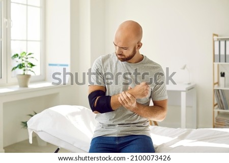 Sick young man sit on couch in hospital have appointment with surgeon about injury. Pain in elbow, ache in arm. Unhealthy ill guy with elastic bandage splint on cubit in clinic. Rehabilitation. Royalty-Free Stock Photo #2100073426