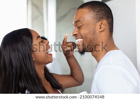Closeup Shot Of Happy African American Couple Having Fun In Bathroom, Cheerful Black Woman Applying Moisturising Cream On Husband's Face, Playfully Touching His Nose And Laughing Together Royalty-Free Stock Photo #2100074548